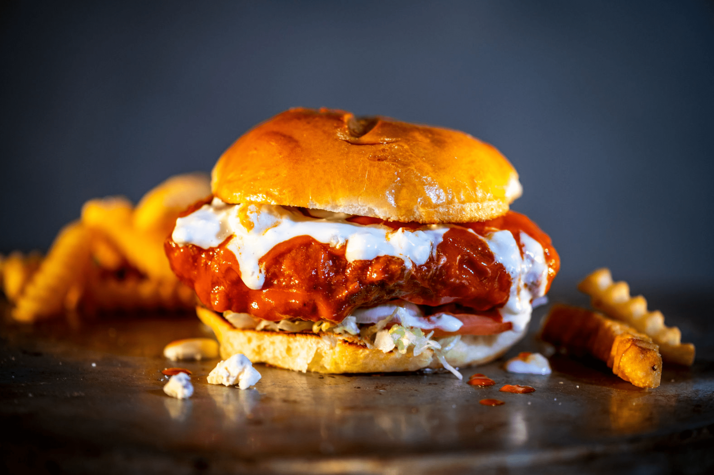 A buffalo chicken sandwich dripping in buffalo sauce and blue cheese dressing surrounded by crinkle cut fries