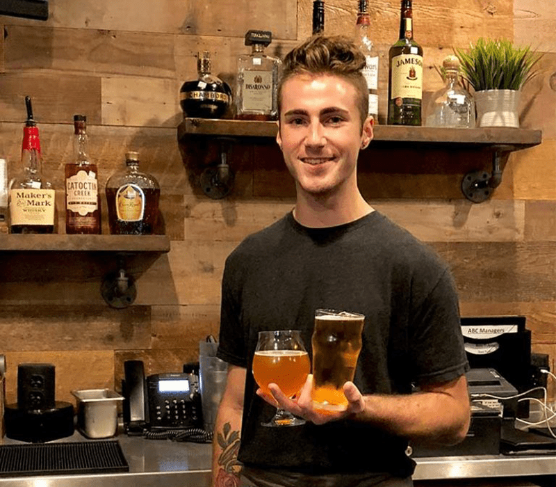 A man holding two different beers in one hand while standing in front of a bar